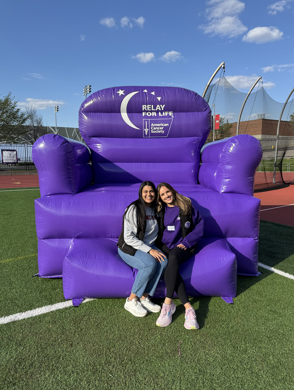 Isha and a friend. sit on a large, inflatable, purple chair that says "relay for life" 