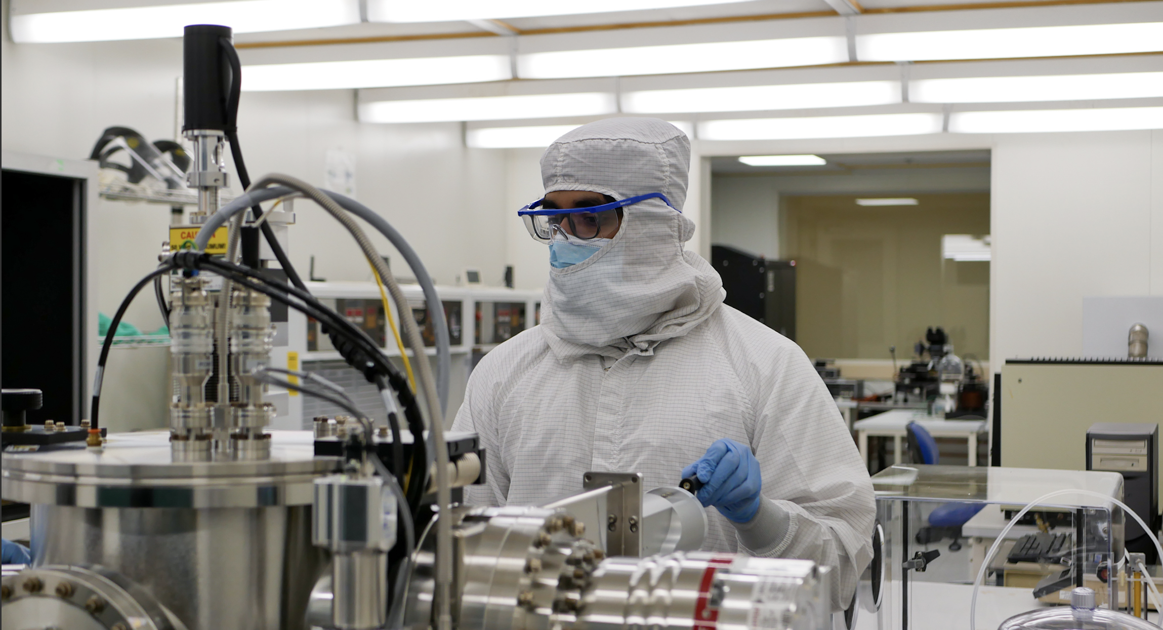 A student covered from head to toe in protective clothing works with stainless steel equipment in a cleanroom.