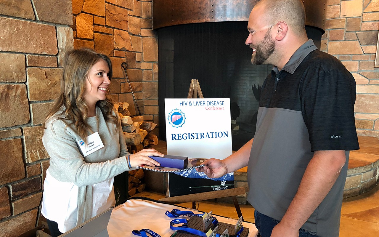 Woman greets male conference attendee at registration table, handing him a program. name badge and gift.