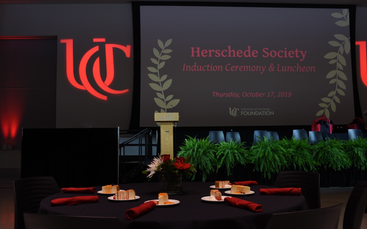 Dark room with UC logo projected on wall. Banquet tables and stage indicate a formal event.