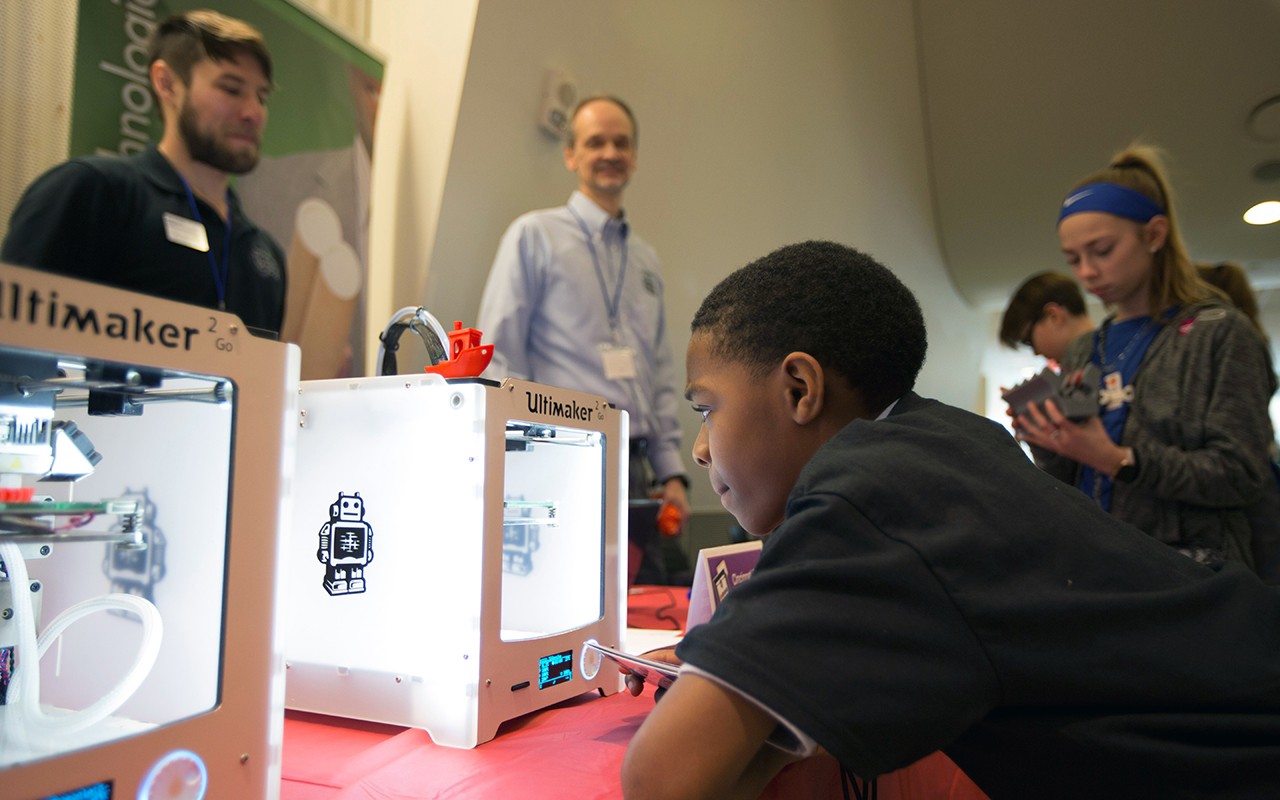 Middle school students look at 3D printer with teachers watching.