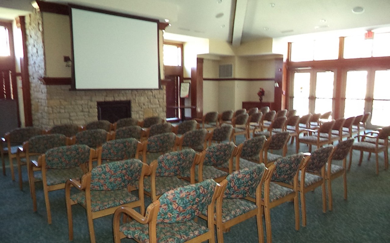 Well-lit meeting room with chairs in rows, lecture style; a projector screen is at the front of the room, above a fireplace.