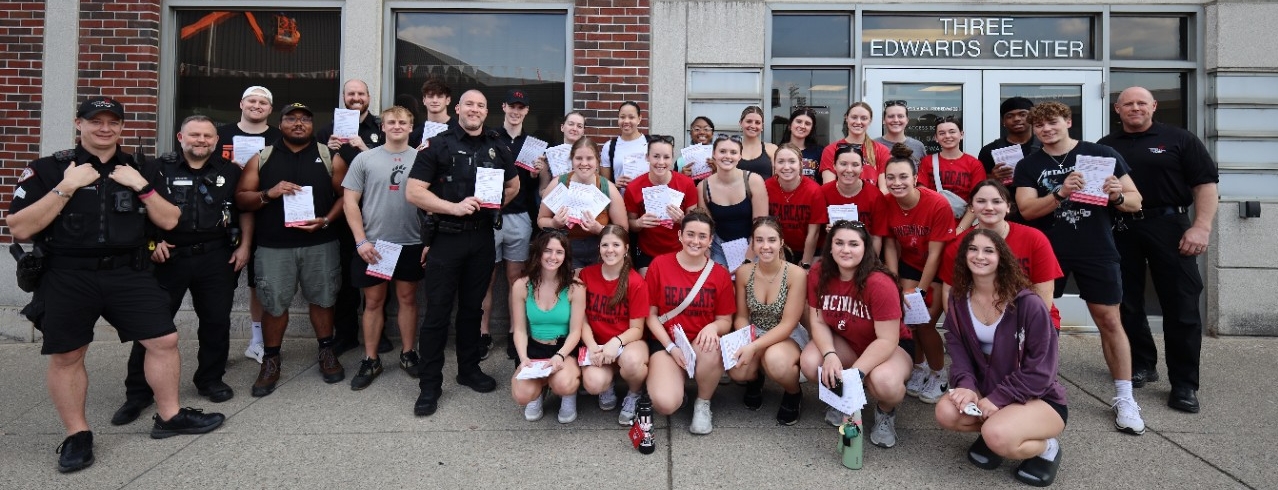 A group of students pose in front of Three Edwards Center with some UCPD Officers holding flyers with burglary prevention information.