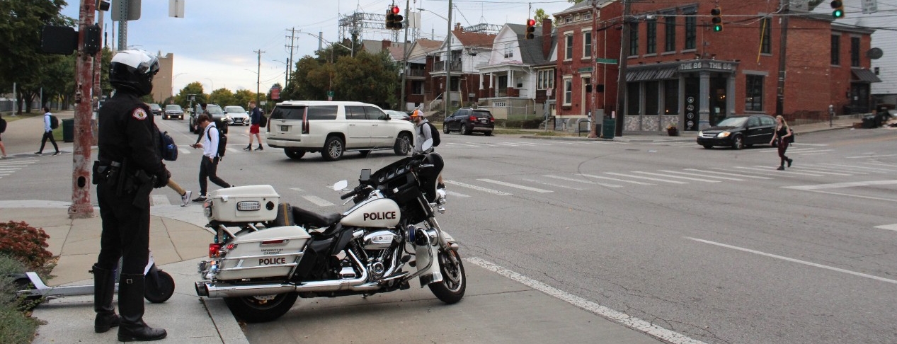 A police officer stands behind a police motorcycle and watches students cross a street.