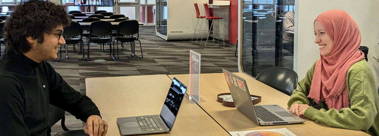 Two individuals engaged in career coaching at a table in the career studio, each using a laptop. One person is wearing a hijab. There are whiteboards and red walls visible in the background.