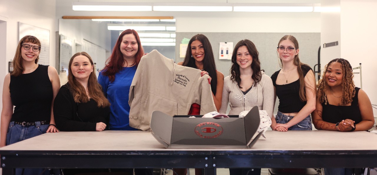   Seven individuals standing behind a table in a workshop setting at the University of Cincinnati, smiling at the camera, with a sewing machine and a textile product displayed on the table.
