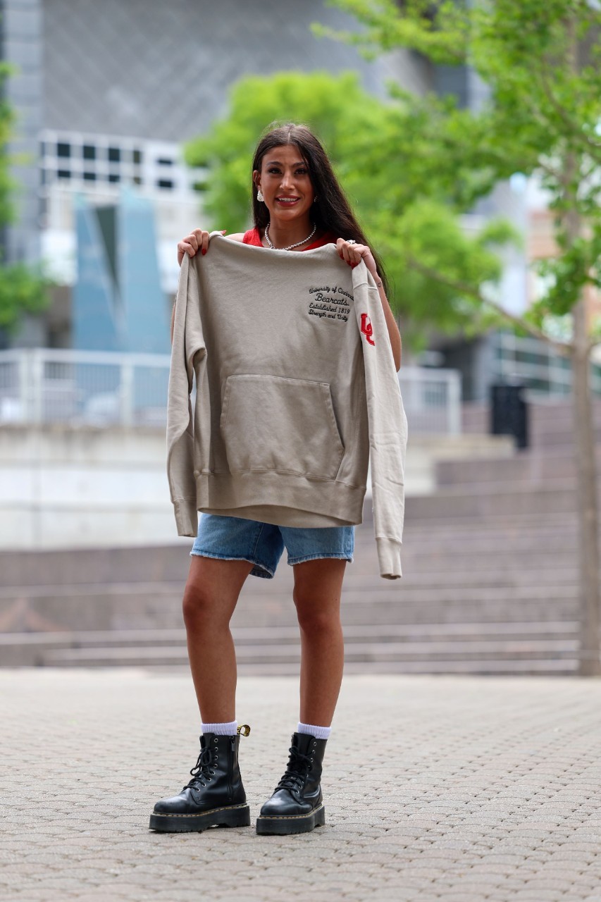 Person holding a UC sweatshirt, standing outdoors with a modern building in the background.