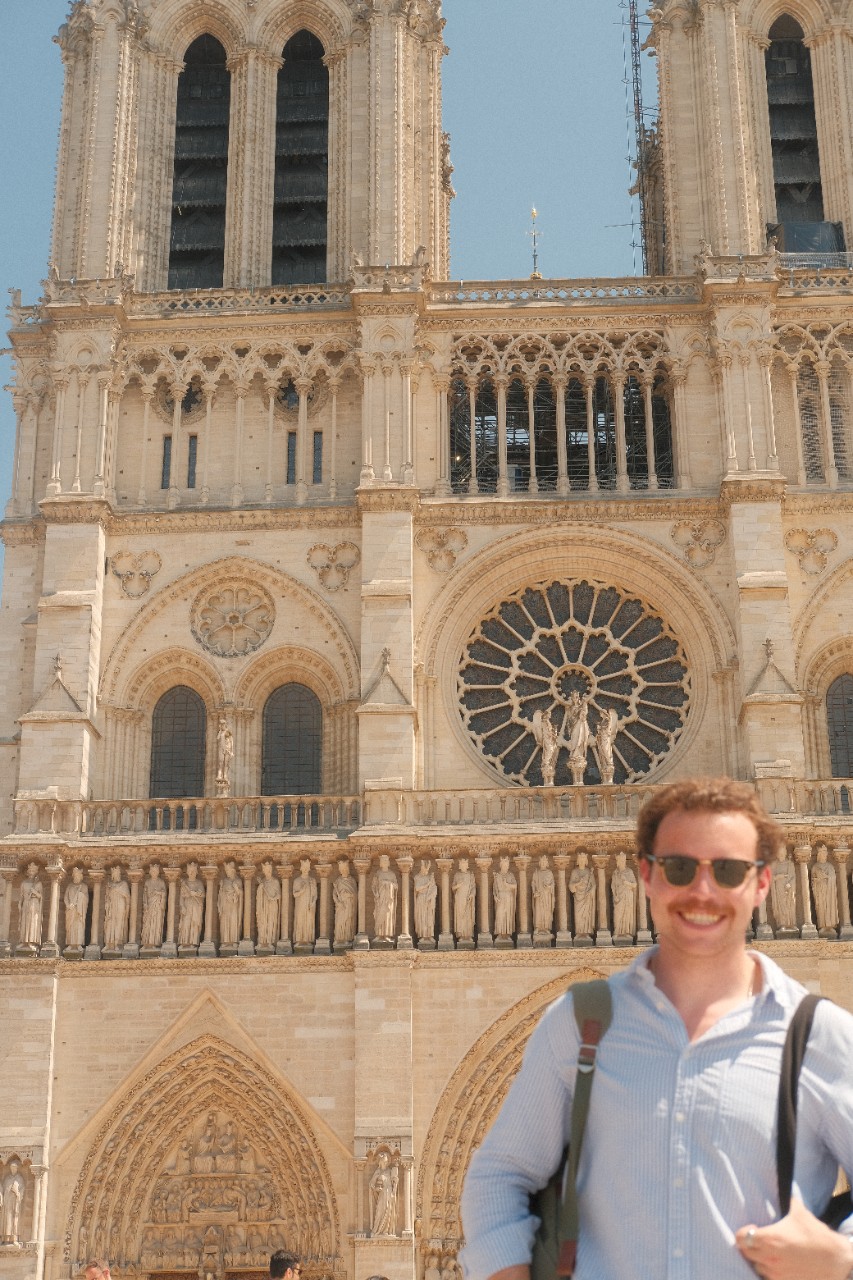 A person smiling in front of the Notre-Dame Cathedral, showcasing the detailed facade and rose window. 