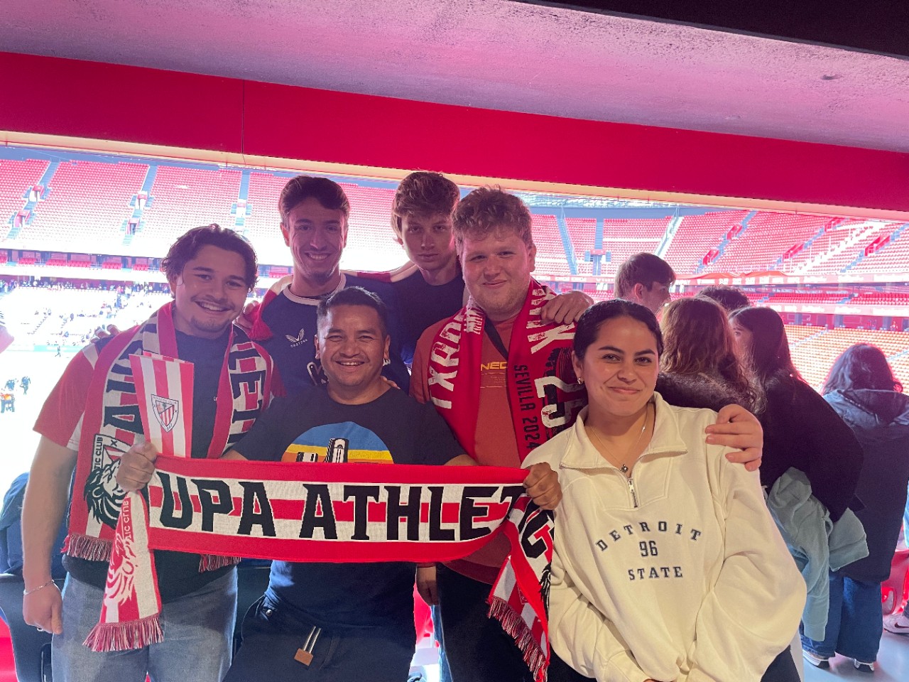 Group of seven soccer fans holding a "UPA Athletic" scarf, smiling at the camera in a stadium. 
