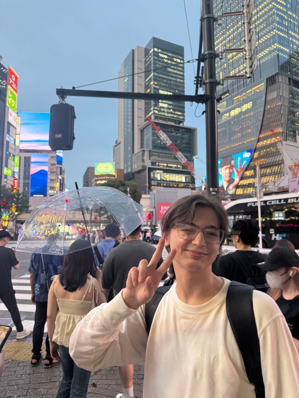 A person making a peace sign in front of the bustling Shibuya Crossing in Tokyo during dusk. They stand in front of a transparent umbrella and wear glasses and a light-colored sweater.