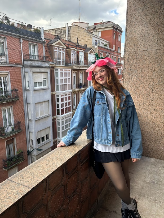 Person smiling, wearing a denim jacket and a pink hat, standing on a balcony with urban buildings in the background. 