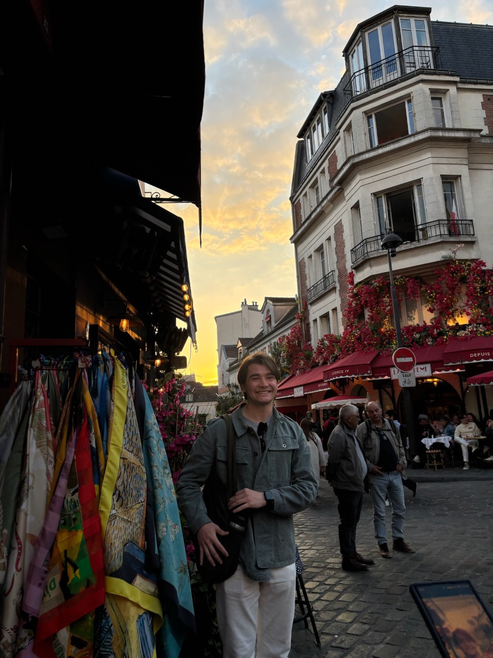 Person smiling in a bustling street market at sunset, with colorful textiles on display and a picturesque building in the background. 