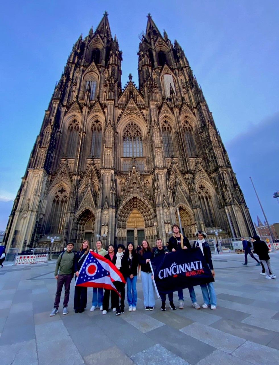 Group of people holding a "Cincinnati Bearcats" banner and flag in front of the Cologne Cathedral