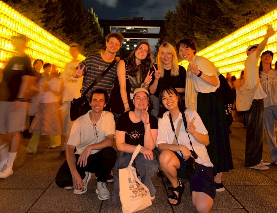 Group of people smiling at the camera during a nighttime outdoor event, with rows of illuminated lanterns on either side. 