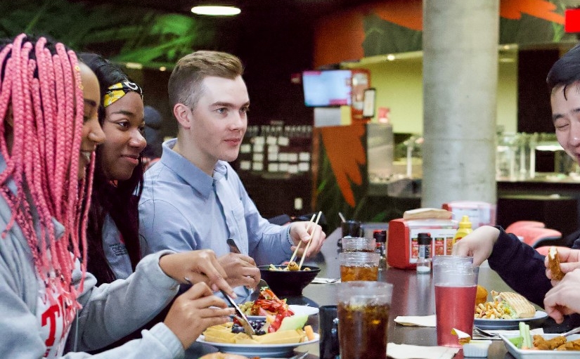 Students sit eating at several tables in the dining center. Two serving stations are in the background