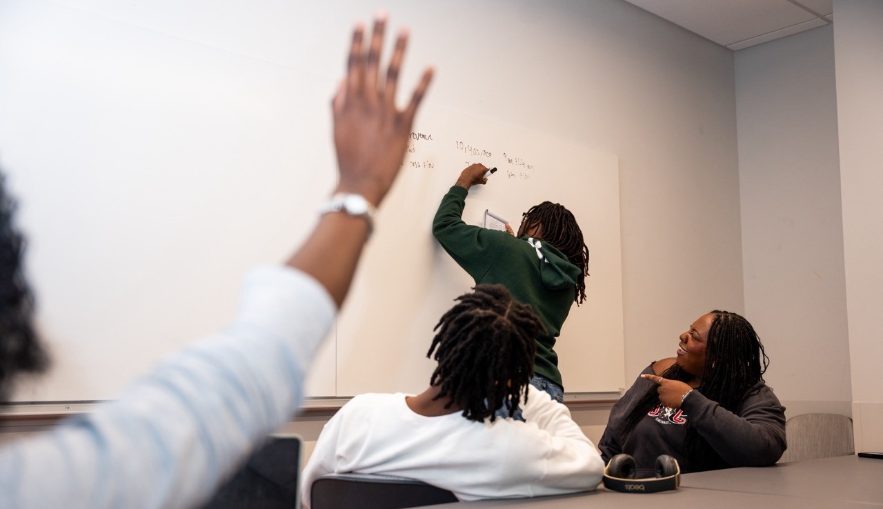 Student raising hand in a class while another student writes on board
