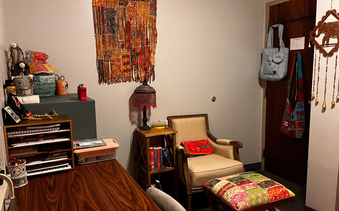 Desk with orange and yellow accessories, a soft chair with a patchwork ottoman, and a colorful tapestry on the wall