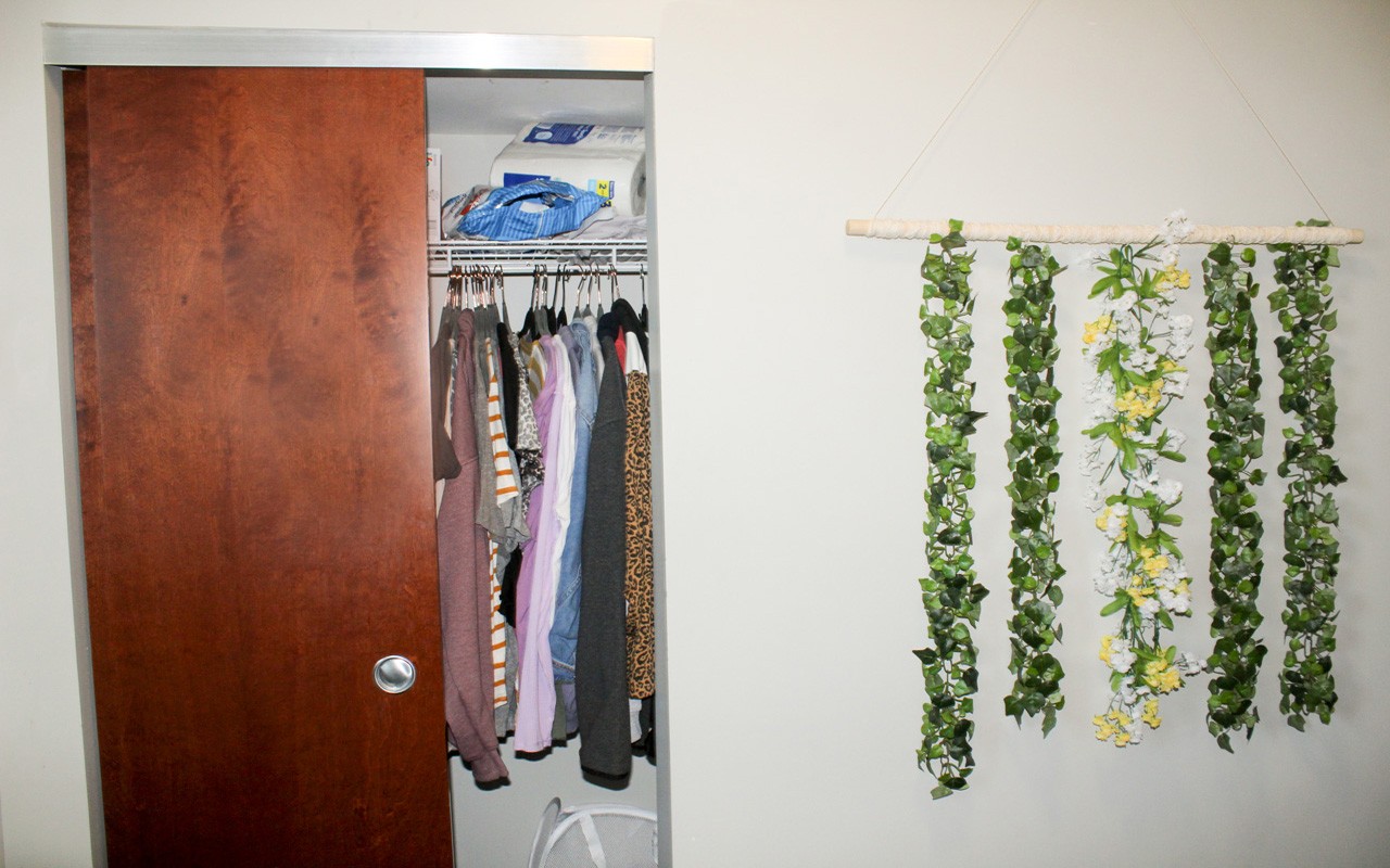 Open closet in a single room in a Stratford Heights suite. There is a rack with clothes hanging from it and other other items on the shelf above.
