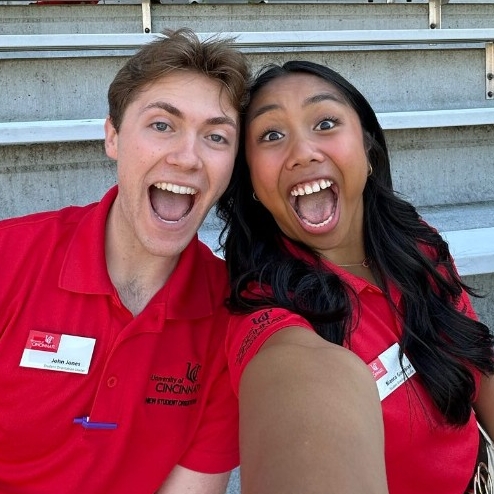 two student orientation leaders with big smiles pose for a selfie with stadium bleachers in the background.