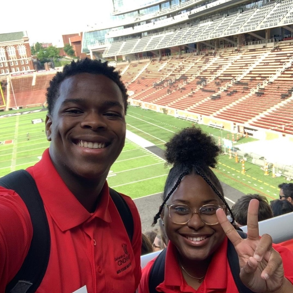 two students wearing UC polos stand in front of Nippert Stadium during orientation. They are both looking at the camera and smiling with one holding up a peace sign.