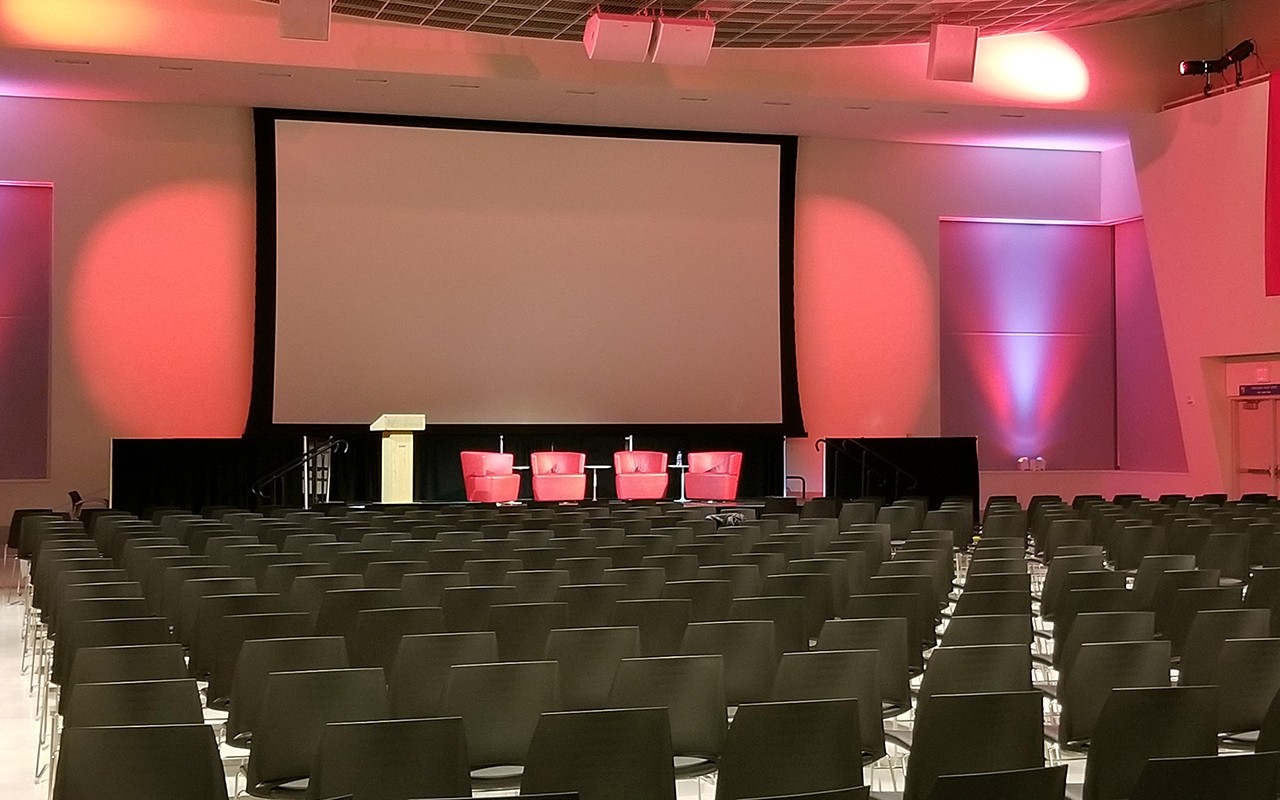 Large meeeting room with many rows of chairs facing a large projector screen. On the stage is a podium and several chairs. There is dramatic lighting on the walls and ceiling around the screen,