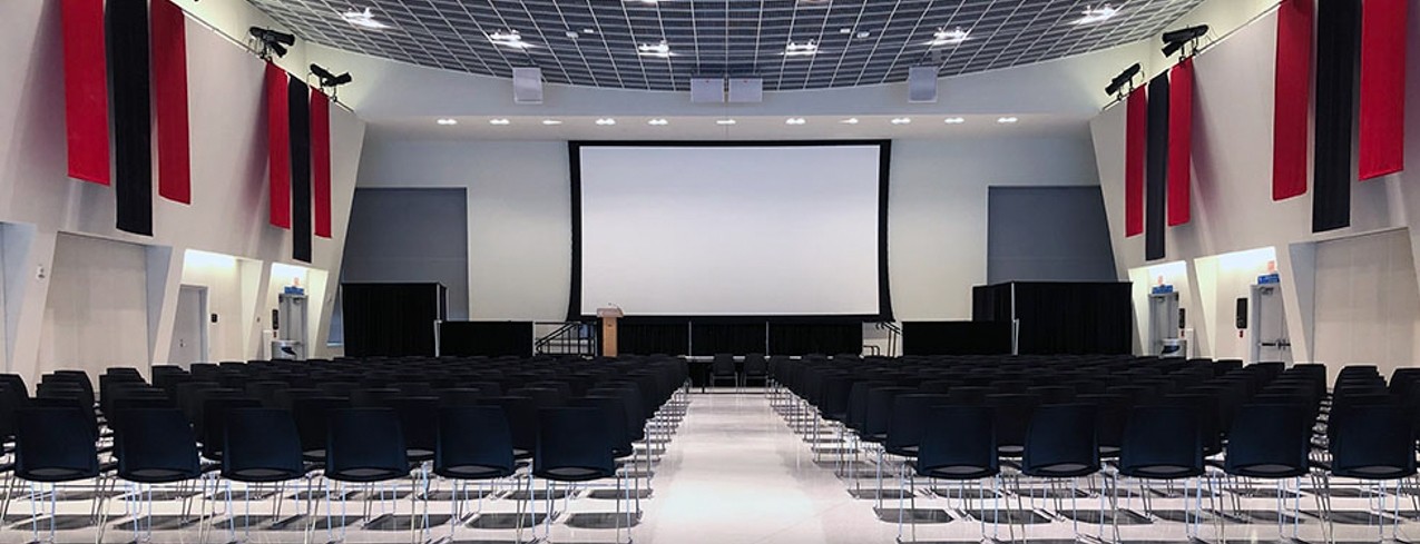Chairs in rows, lecture style with large screen at front of room; banners hang from high walls.