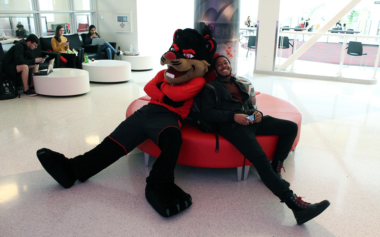 The Bearcat mascot lounges with an African American student.on a settee in the TUC main lobby