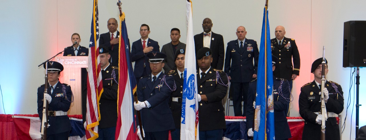UC Army ROTC Color Guard stands at attention in front of the dais, where university and military leaders look on during the singing of the National Anthem.
