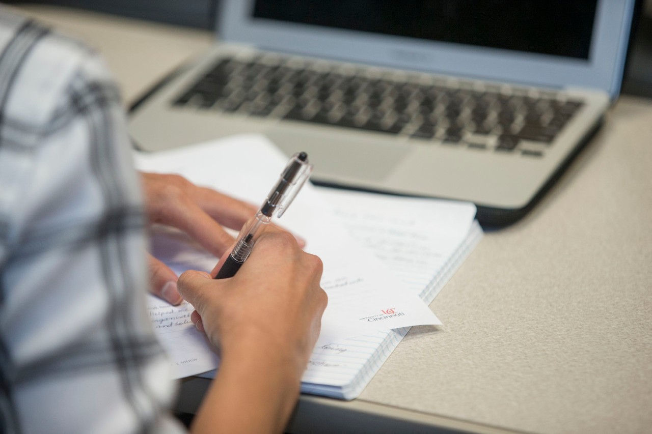 a student sits at a desk a pen in their hand and a notebook and laptop in front of them