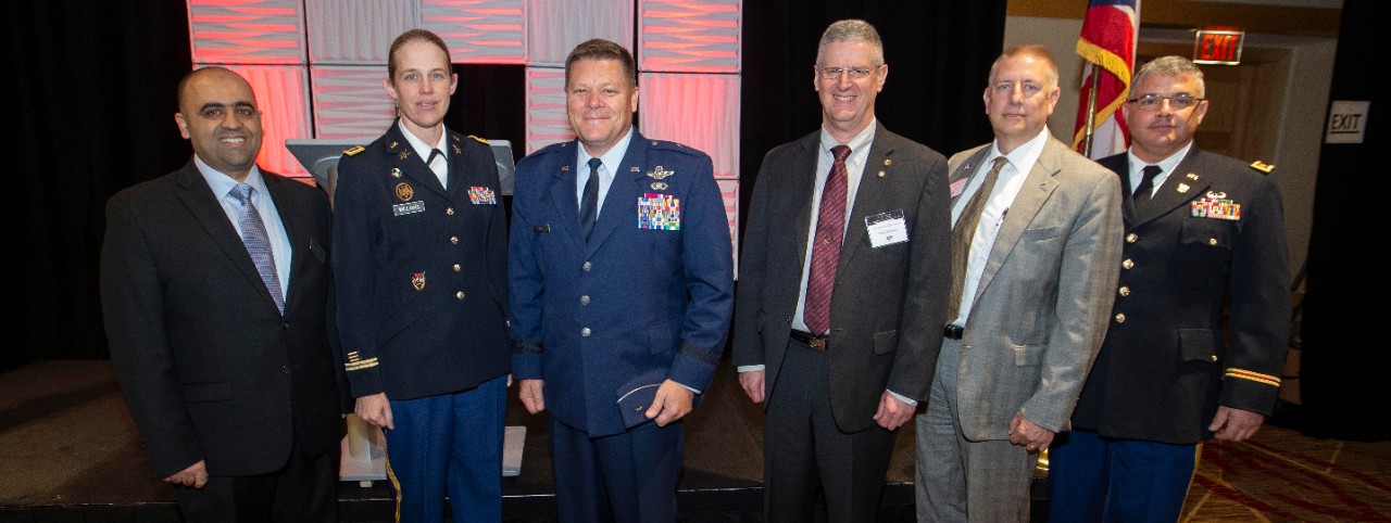 Three men in suits and two men and a woman in military uniforms posing for a photo