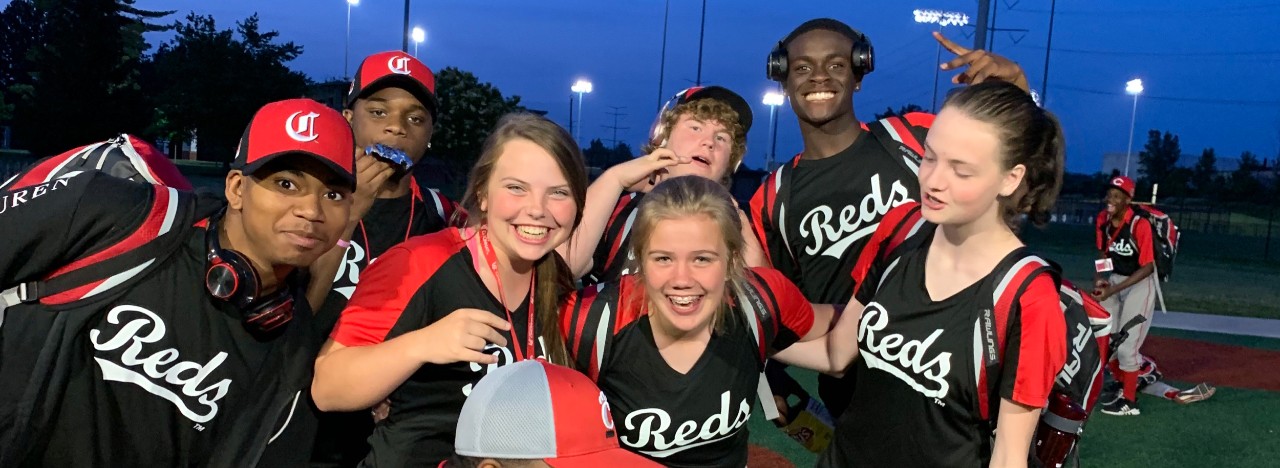 Seven high school-age baseball players stand together wearing Cincinnati Reds uniforms