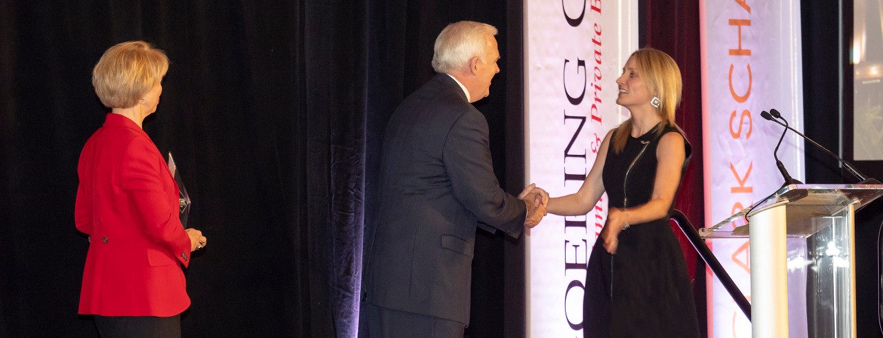 A man in a suit shakes hands with a woman in a black dress near a podium with banners in the background. Another woman in a red coat looks on from the left