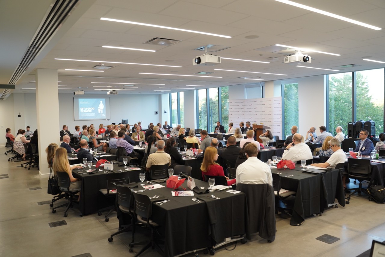 Nearly 75 attendees sit at square tables in a large room looking at a panel of five speakers at the front
