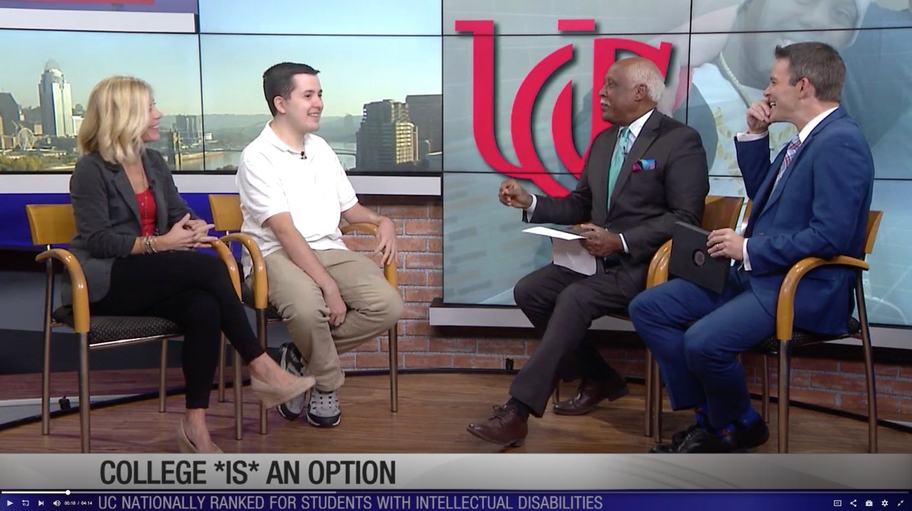 Two men in suits on the right talk to a young man in a white polo shirt and a woman dressed in business casual attire