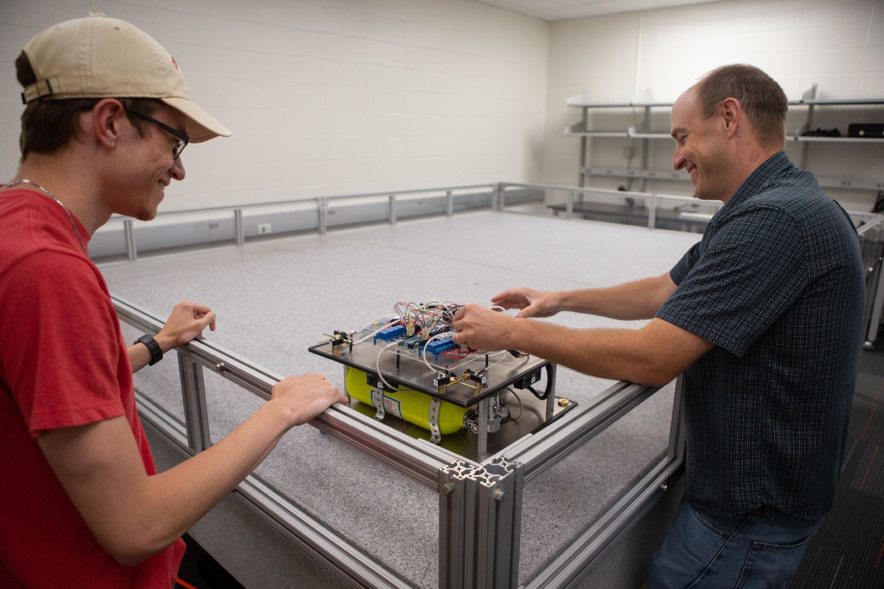 Two students work on a shoebox-sized robot that sits on a metal table surrounded by bumpers.
