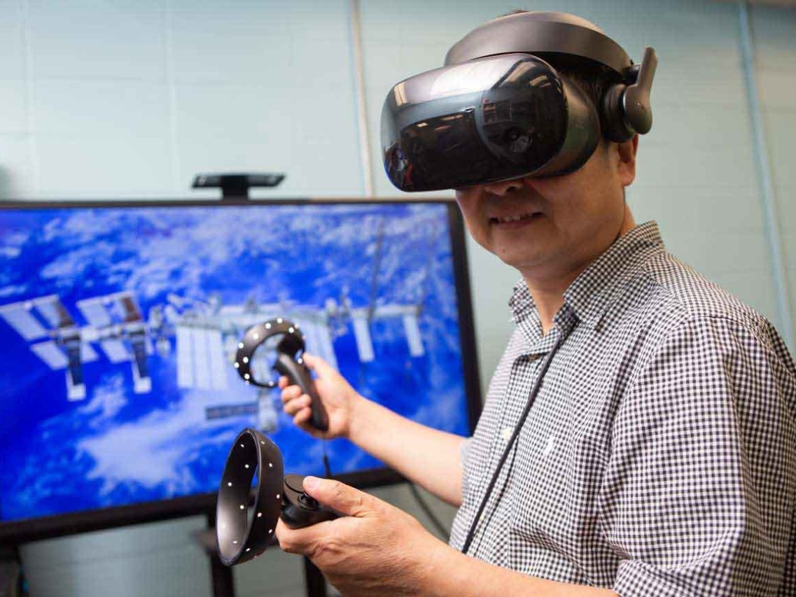 Ou Ma wears virtual reality headset in front of a screen depicting the International Space Station.