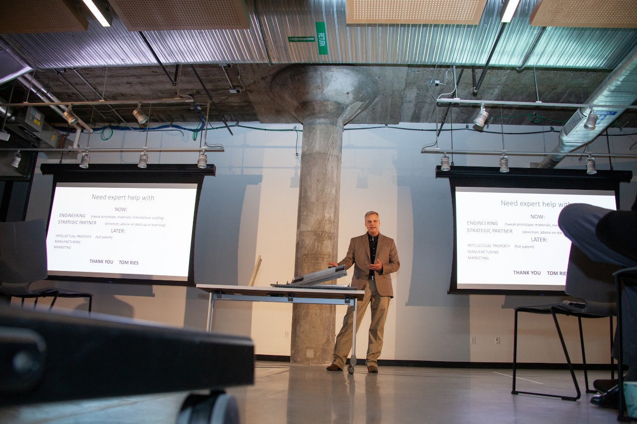 A wide shot of a man in a brown suit standing behind a table with two large video screens behind him to either side