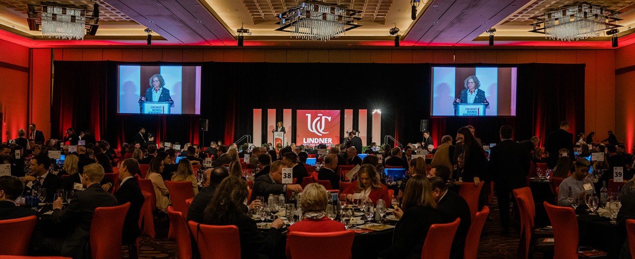 A woman stands and speaks at a podium on a stage in front a large ballroom with hundreds of people seated at round tables