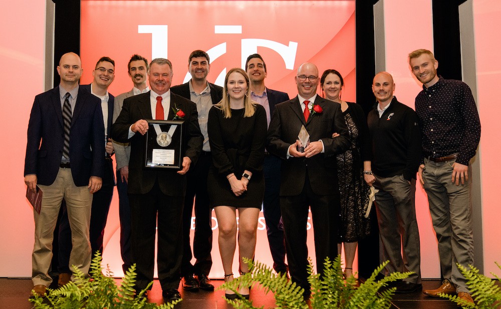 Eleven employees from award-winning company Enerfab stand and smile on a stage with a UC backdrop