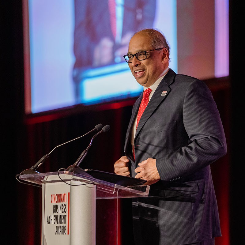 UC President Neville Pinto stands and speaks from a clear podium on a stage, wearing a dark suit and a red tie