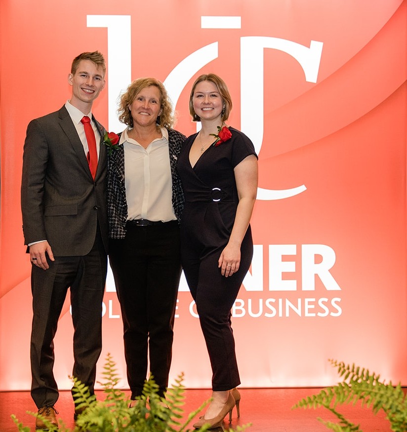 A young man stands next to Dean Marianne Lewis and a young woman on a stage with UC backdrop