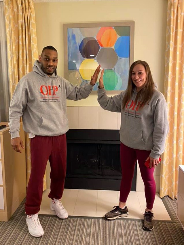 A man in a grey sweatshirt and maroon jogging pants gives a high five at a distance to a woman wearing the same grey sweatshirt and maroon leggings