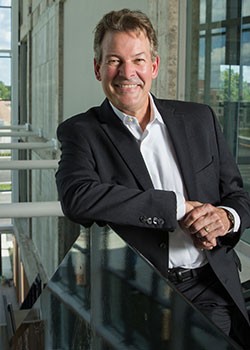 A man in a sport jacket and button down shirt smiles while leaning against the top of a stairwell