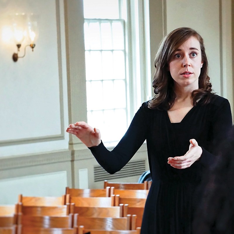 A woman conducts a choir in a rehearsal room