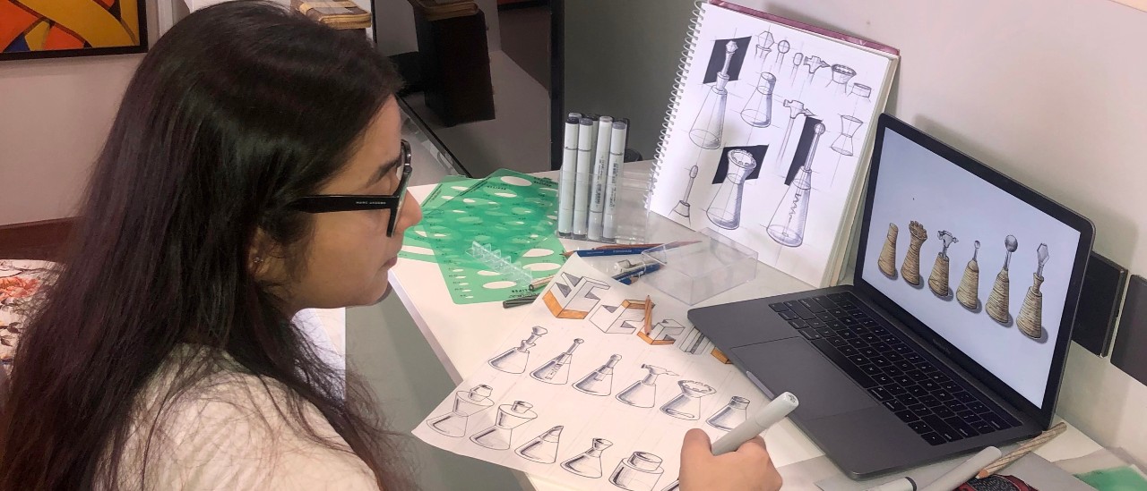 UC's Reeha Das sits at computer while drawing designs on paper.