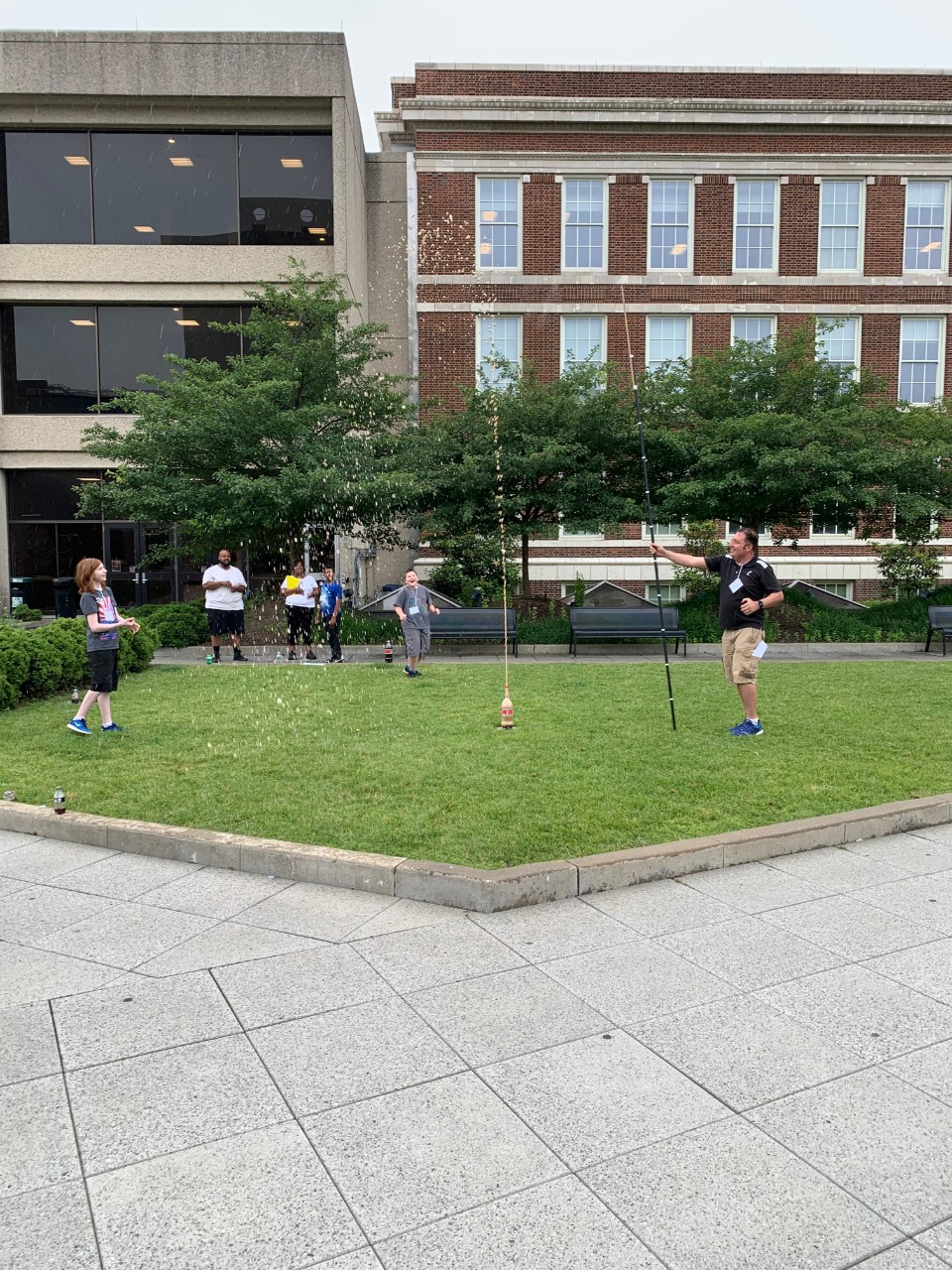 Middle school students and parents stand outside a building on a lawn, watching several cola bottles explode with foam