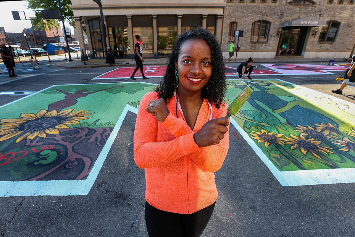 UC's Adoria Maxberry stands holding a paint brush in front of her BLM mural on Plum Street.