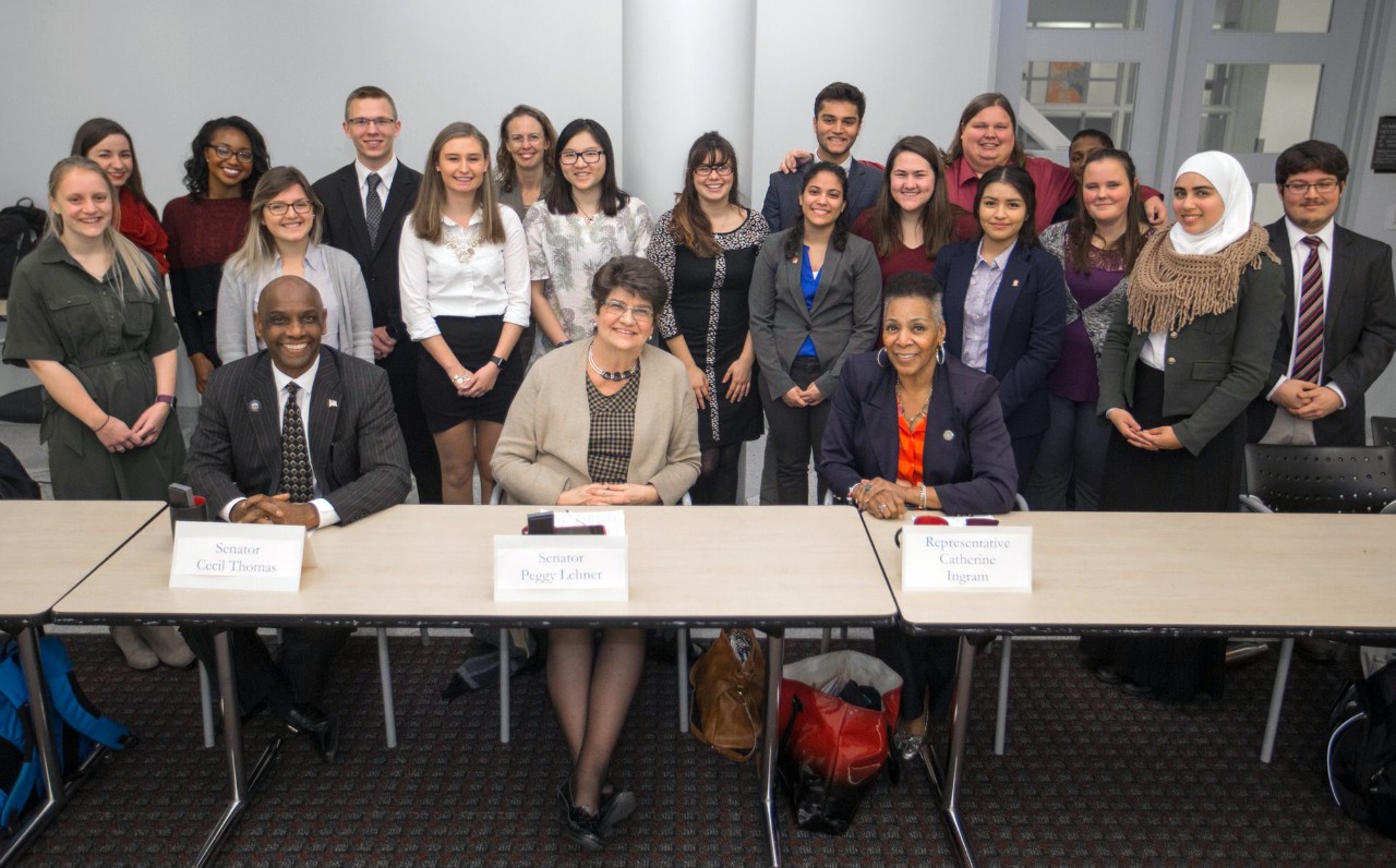 a group of students stand behind three seated people at a desk