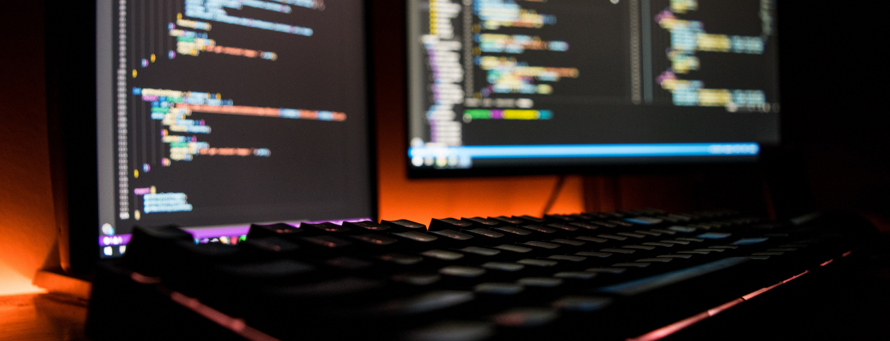 a desktop with keyboard in the foreground and two monitors in the background displaying code