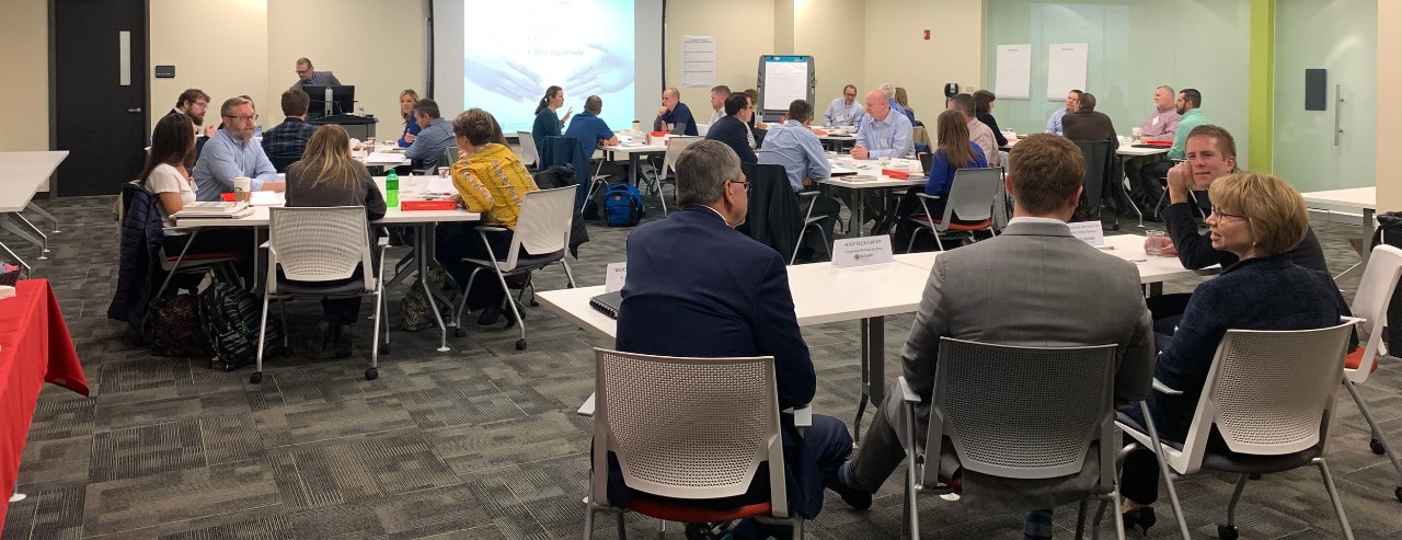 a classroom full of business people sitting at multiple tables with a projection screen at the front of the class